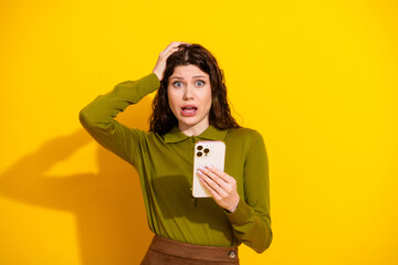 Surprised young woman holding a smartphone and expressing emotions on a yellow background, wearing...