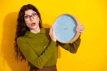 Young woman in khaki shirt holding blue clock against vibrant yellow background expressing surprise...