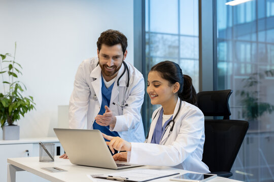 Two doctors, a man and a woman, collaborate at a desk in a modern office. They are looking at a laptop screen.