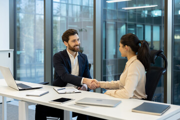 A smiling man and woman shake hands at a desk in an office setting, possibly during a business meeting or interview.