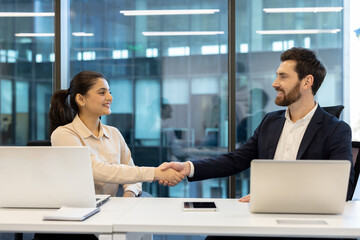 A business meeting is in progress, with two professionals shaking hands to finalize an agreement in a modern office setting.