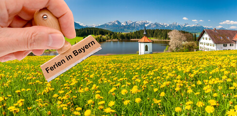 Vor einer idyllischen Landschaft in Bayern wird ein Holzstempel gehalten mit der Aufschrift: Ferien...