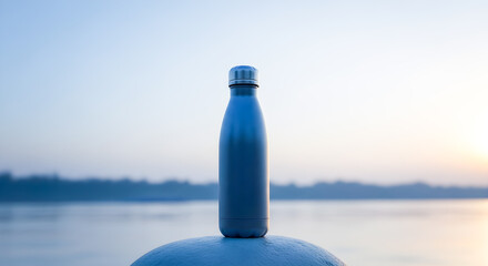 Stylish reusable water bottle is standing on a post near the water at dawn, promoting healthy...