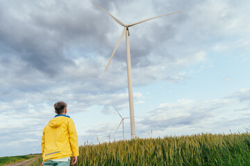 Back view of teen boy in yellow jacket standing in wheat field, looking at large wind turbine under...
