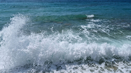 A powerful wave crashes into the shore, creating a vibrant splash of white foam against the deep blue and turquoise hues of the sea
