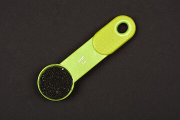 A vibrant green 1 teaspoon (5 ml) measuring spoon filled with black Hawaiian salt is centrally positioned on a dark background