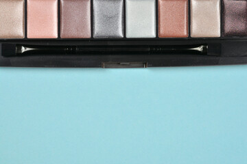Shimmering eyeshadow palette with various metallic shades of silver, pink, and brown, including a small applicator brush, on a light blue background