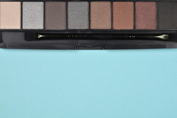 Dark eyeshadow palette with various shades of brown and gray, including a small applicator brush, on a light blue background.