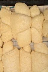 A close-up overhead shot shows rows of raw, pale yellow mini croissants sprinkled with sesame seeds on a grid surface