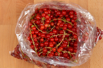 A top-down view of vibrant red currants with their green stems, freshly packed in a clear plastic bag on a wooden surface