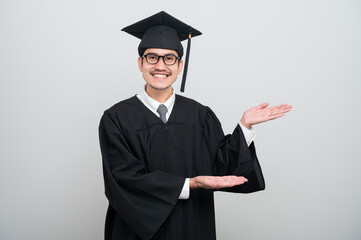 A young man in a graduation gown and cap smiles at the camera, striking a confident pose to...