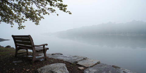 A lone, weathered wooden bench placed on the edge of a foggy lakeshore, surrounded by soft gray tones. The still water reflects