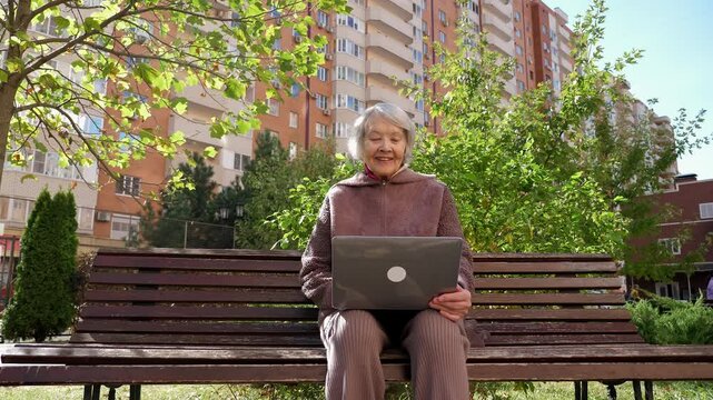 A stabilized image of an elderly modern woman with a laptop in the park on a bench. An octogenarian grandmother with a laptop in the courtyard of a multi-storey residential complex in autumn.