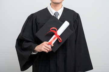 A graduate proudly holds up a rolled diploma tied with a red ribbon and a graduation cap, striking...