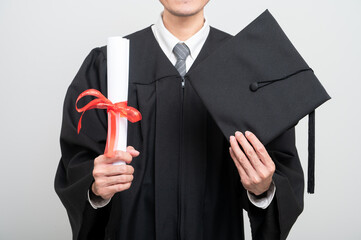 A graduate proudly holds up a rolled diploma tied with a red ribbon and a graduation cap, striking...