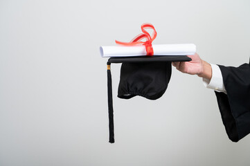 A graduate proudly holds up a rolled diploma tied with a red ribbon and a graduation cap, striking...