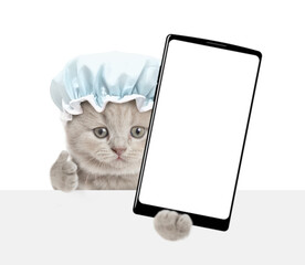 Funny tiny kitten wearing shower cap holds big smartphone with white blank screen in it paw above...