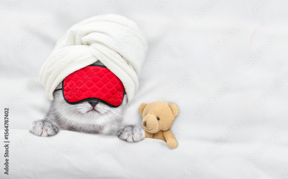 Poster cute tiny kitten with towel on it head and mask for sleep relaxing with toy bear on a bed at home. t