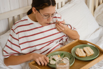 Teen girl wearing eyeglasses having breakfast sitting in bed, drinking tea, enjoying her morning at...