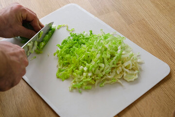 Handsome bearded man is preparing green fresh vegan salad in the kitchen at home. Male cutting vegetables, lettuce with knife on wooden chopping board, cooking vegetarian meal. Tasty healthy diet food