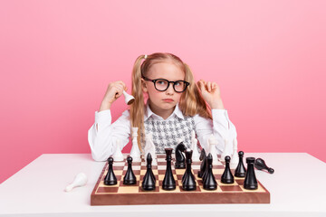 Focused young student analyzing a chess game on a wooden board against a pink background in a...