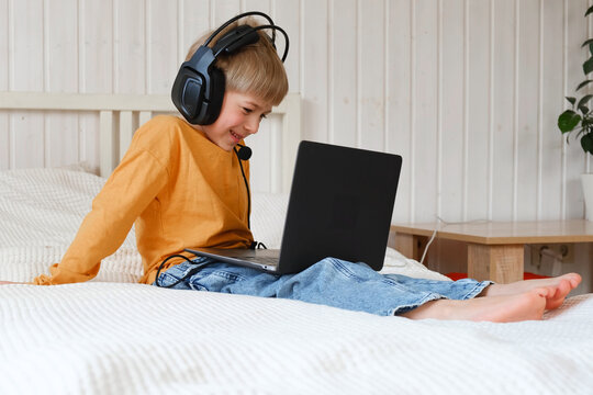 Little smiling 6 years old boy using a laptop with headphones. Child having video call with friends, granny at home in bed. Kid looking at display and waving hand. Childhood leisure activity concept