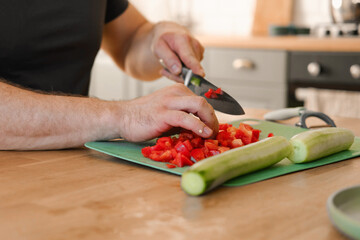 Man preparing fresh vegetables in modern kitchen, organic ingredients. Healthy meal. Cooking Food At Home. Male slicing red pepper and green cucumber on a wooden cutting board. Nutritionist, culinary