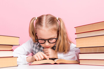 Young schoolgirl in glasses studying with book on pink background, symbolizing education and...