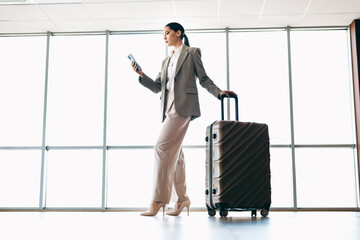 Professional woman in formalwear with luggage at modern airport terminal holding phone,...