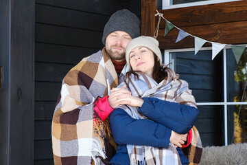 Couple relaxing on a patio wrapped on checkered blankets. Relaxed beautiful woman with a handsome man sitting on wooden cottage terrace. Winter resort. Happy loving couple enjoying leisure, embracing