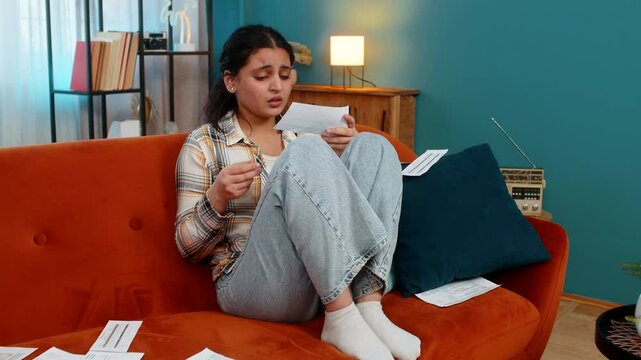 Indian young woman sitting on sofa looking at bills, receipts, praying with eyes upward, sincerely asking for help. Arabian girl experiencing hope desperation, emotionally praying hoping for a miracle