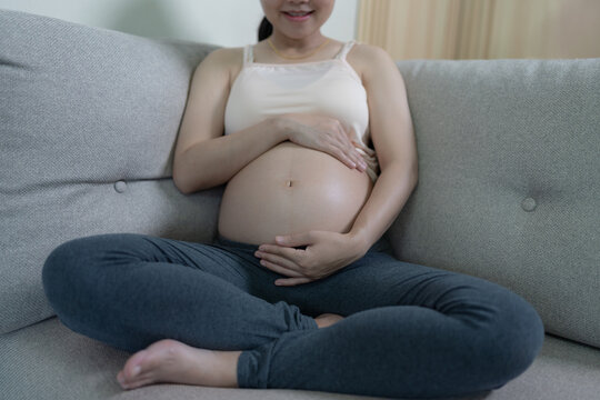 A smiling pregnant woman sits cross-legged on a sofa, gently cradling her bare belly with both hands, symbolizing calmness and maternal joy.