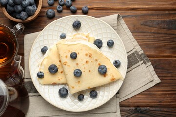 Tasty crepes with cream, blueberries and tea on wooden table, flat lay