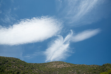 Wispy clouds stretch across a vibrant blue sky, casting shadows on lush green hills below, capturing a serene and tranquil landscape