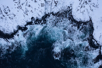 Aerial perspective showcasing powerful ocean waves crashing against a rugged, snow covered...