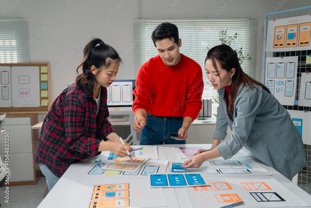 Poster three asian ux designers collaborating on a mobile application prototype, sketching ideas on a large