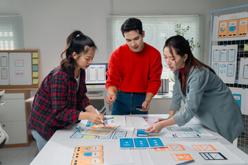 Three Asian UX designers collaborating on a mobile application prototype, sketching ideas on a large desk in their modern office, fostering creativity and teamwork