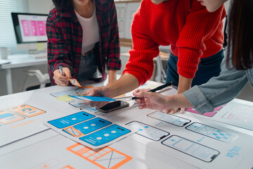 Three Asian ux designers are discussing and improving the design of a mobile application during a working meeting, placing different pieces of paper and sticky notes on the table