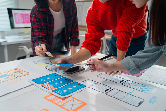 Three Asian ux designers are discussing and improving the design of a mobile application during a working meeting, placing different pieces of paper and sticky notes on the table