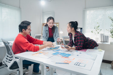 Group of young Asian UI/UX web designers collaborating in a modern office, analyzing and reviewing mobile app development on paper layout prototypes