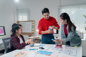 Three Asian UX designers collaborating on a new mobile app interface in a modern office, analyzing...