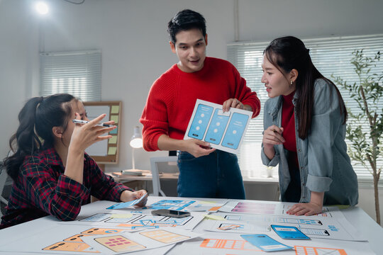 Diverse team of young designers engaged in a collaborative mobile app development meeting, analyzing wireframes and discussing user interface improvements in a modern office setting - Powered by Adobe
