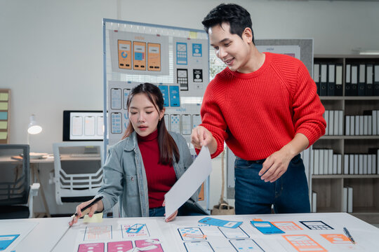 Two young Asian UX/UI designers collaborating in a modern office, creating mobile app interface wireframes for an innovative application, fostering creativity and teamwork