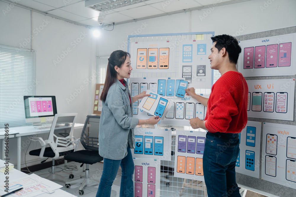 Poster two young asian designers comparing mobile app templates hanging on a board, discussing user interfa