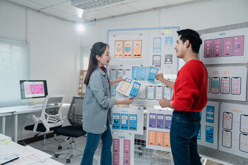 Two young asian designers comparing mobile app templates hanging on a board, discussing user...
