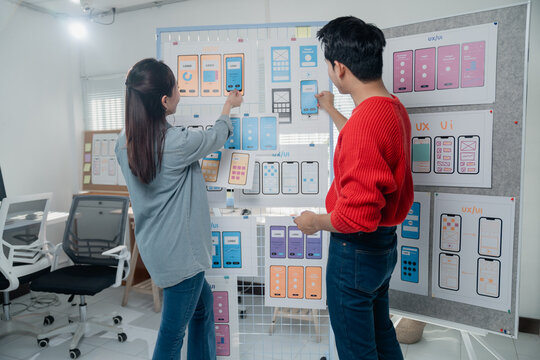 Two UX/UI designers collaborating on app design workflow, arranging printed templates of app screens on a whiteboard in an office setting, fostering creativity and teamwork