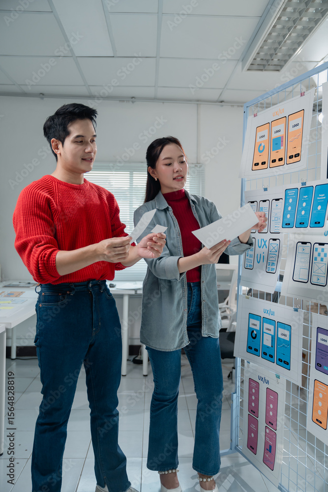 Poster two young ux/ui designers comparing printed mobile app templates on paper sheets while standing by a