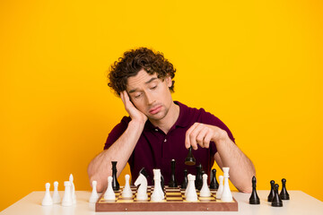 Young man playing chess while showing a reflective expression sitting at a table against a bright...
