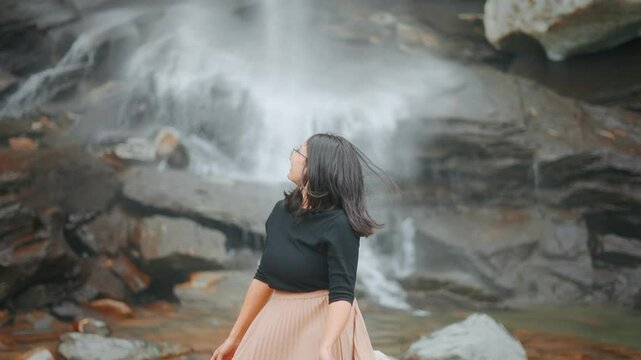 4K Portrait of Indian woman looking at beautiful waterfall in Sural Bhatori, Pangi Valley, India. Tourist exploring hidden gems in mountains. Woman enjoying tranquility by a waterfall