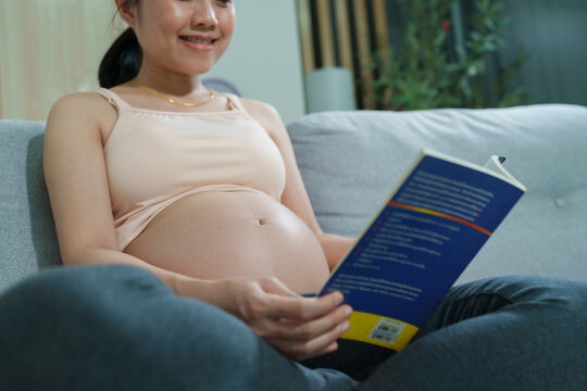 Expecting mother relaxing on a sofa while reading a book, smiling gently with hands on her belly, capturing a peaceful and nurturing moment at home.